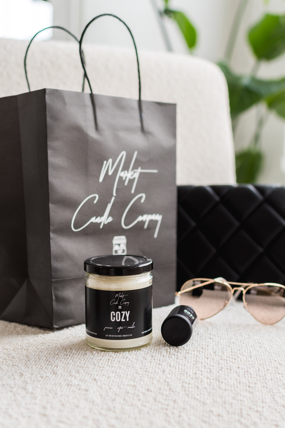 Candle with 'Cozy' label, sunglasses, and a branded gift bag on a textured surface.