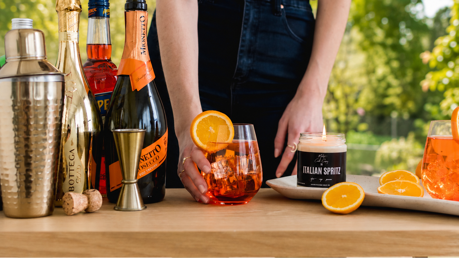 Person standing behind a table with cocktail-making ingredients and a drink, with a blurred outdoor background.