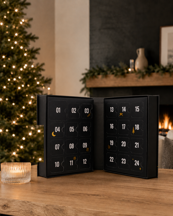 A black 24-door advent calendar stands open on a wooden table in a cozy indoor setting with a lit Christmas tree, fireplace, and candle in the background.