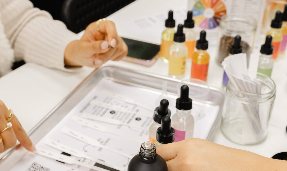 Person holding a bottle oF OIL next to a tray with FRAGRANCE bottles and a color chart on a table.